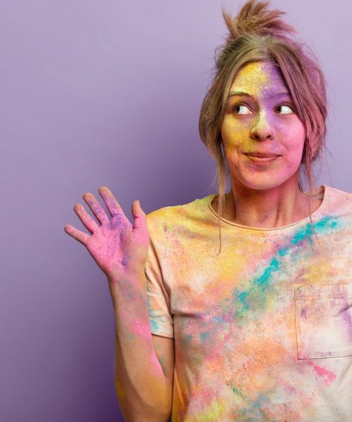 Carefree beautiful female model looks aside with dreamy curious expression, raises palms smeared with colorful powder, involved in Holi festival celebrations, stands over purple wall, blank space